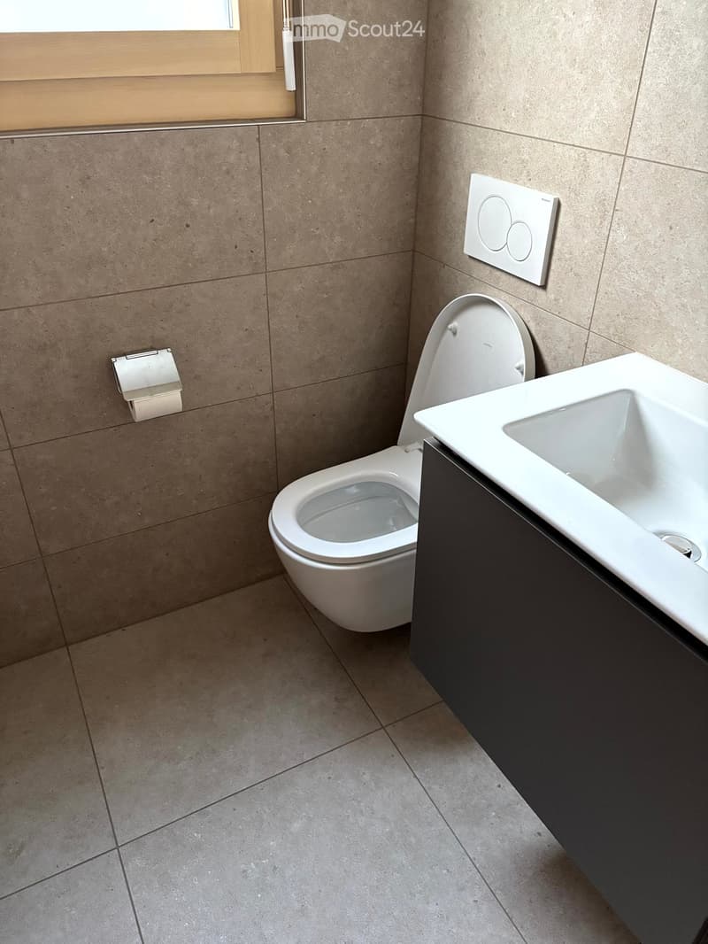 Modern bathroom with beige tiled walls and floor, white toilet, sink with dark cabinet, and a toilet paper holder on the wall.