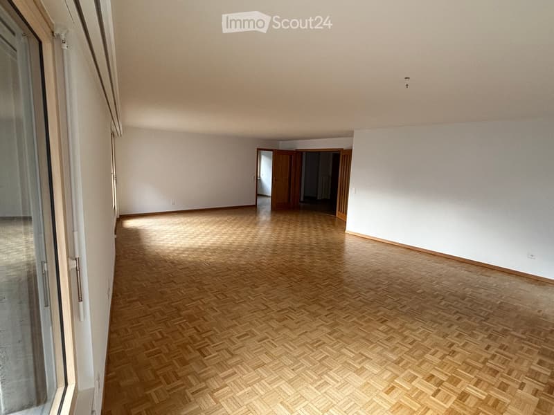 Empty room with wooden herringbone flooring, white walls, and large windows. No furniture or equipment visible.