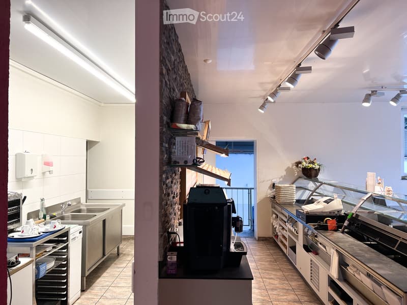 Modern kitchen area with stainless steel appliances, a coffee machine, and a counter with a cash register. Shelves hold bags and boxes, and a flower vase is on display.