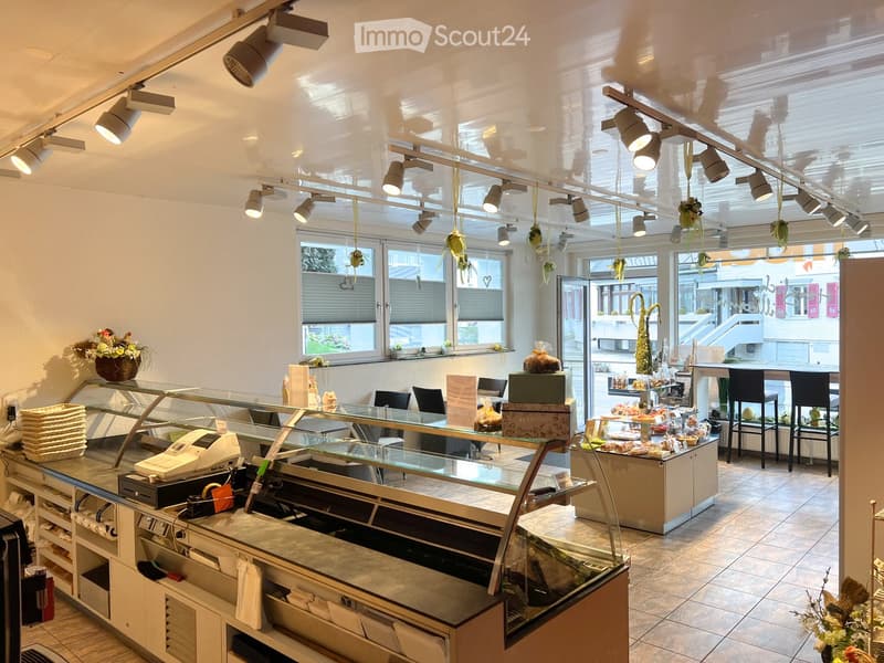 Brightly lit bakery or cafe interior with a display case, cash register, stools, tables, chairs, and decorative flower vases.