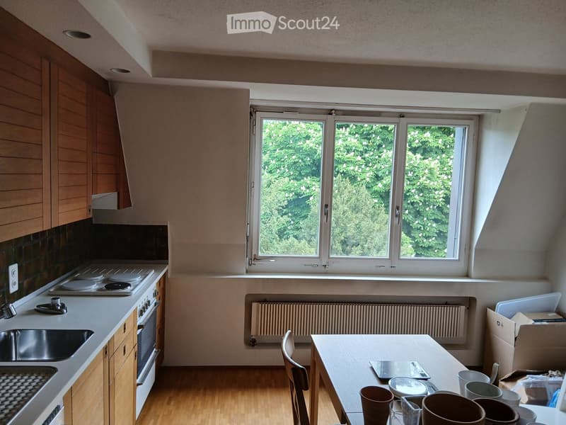 Bright kitchen with wooden cabinets, white countertops, and a large window overlooking greenery. Includes a sink, stove, oven, and a dining table with chairs.