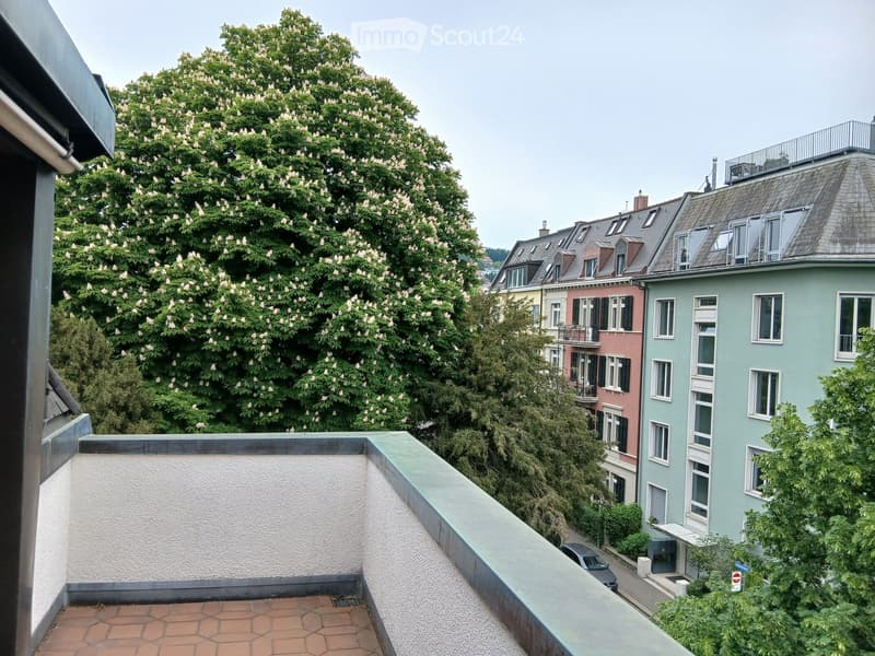 Balcony with a tiled floor and white railing, overlooking a street lined with colorful residential buildings and lush green trees.