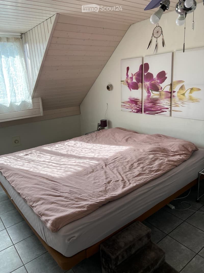 Bedroom. Cozy bedroom with a wooden slanted ceiling, a bed with a pink duvet, a small stool, a wall-mounted lamp, and a large picture frame with floral artwork.