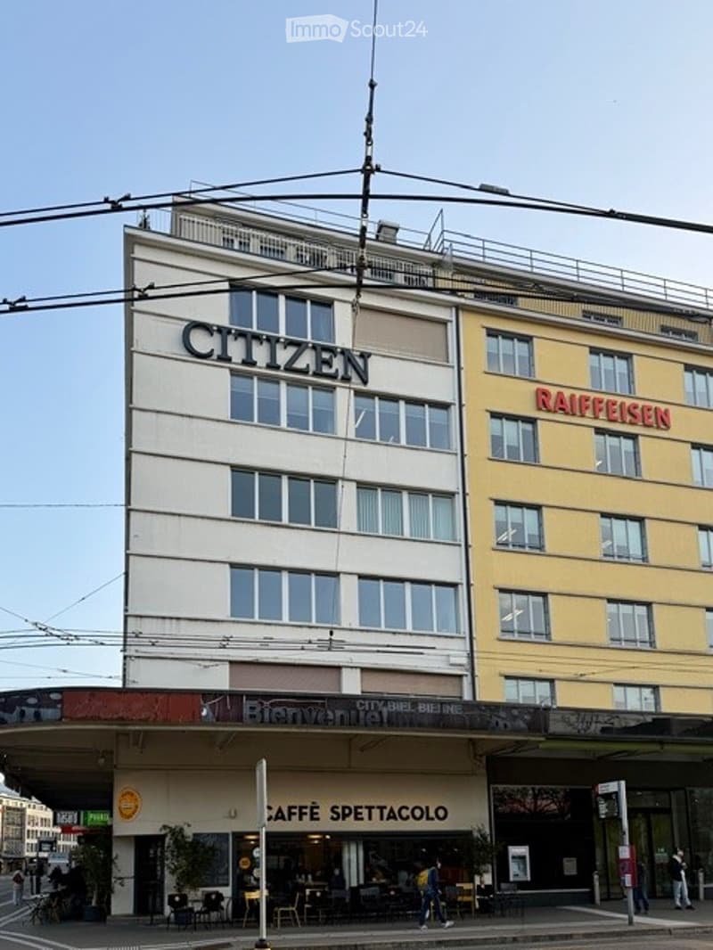 large commercial building, exterior, multiple floors, white and yellow facades, signage reading 'CITIZEN' and 'RAIFFEISEN', several storefronts and entrances, with a cafe entrance reading 'CAFFE SPETTACOLO' in front.