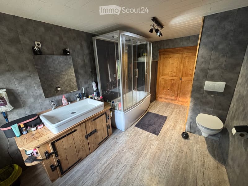 Bathroom. Modern bathroom with a large sink, toilet, bathtub with glass shower enclosure, wooden cabinet, and mirror. The floor is wooden, and the walls are grey.