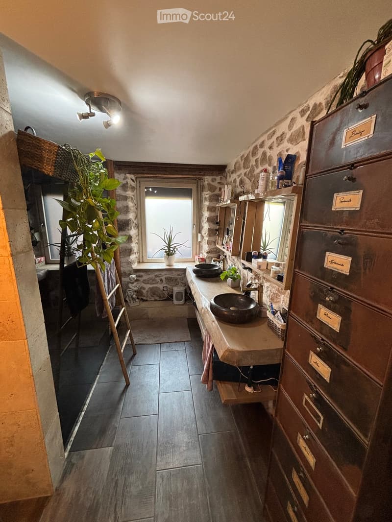 Bathroom. Bathroom with stone walls, dark tiled floor, double sink vanity, mirror, and various toiletries. Features include a window with a small plant on the sill and a tall filing cabinet with labeled drawers.
