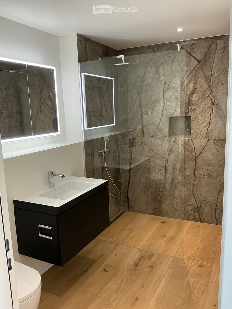Modern bathroom with a sleek black vanity, white sink, chrome faucet, large mirror, and a glass-enclosed shower with marble tiles.
