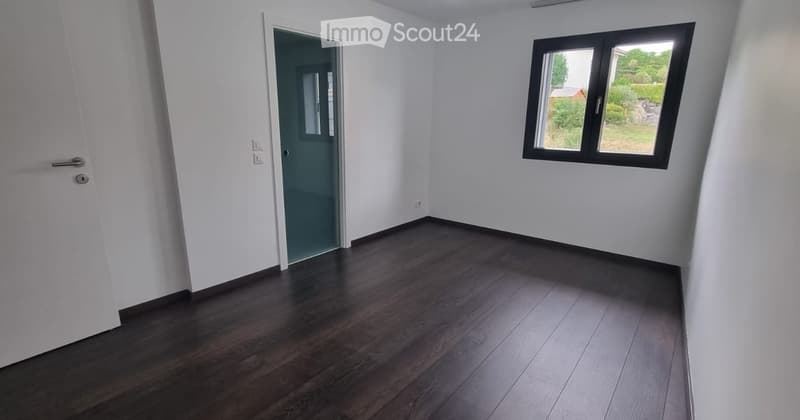 Empty room with white walls, dark wooden flooring, and a single window with a view of greenery. A door leads to another room.