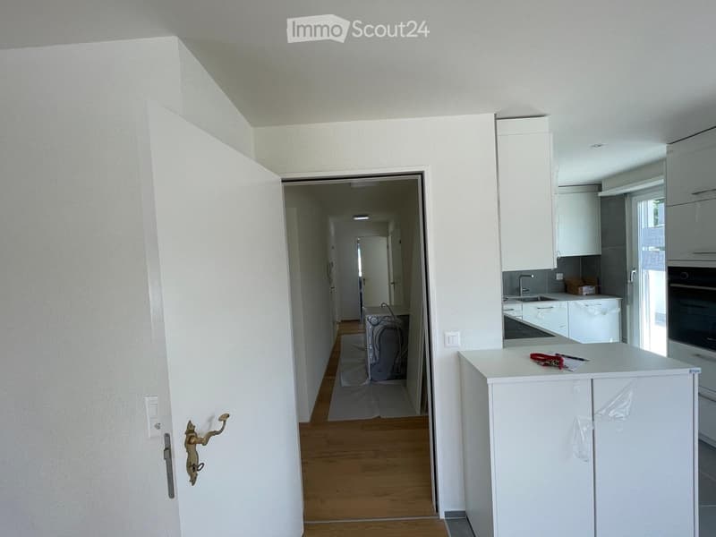 A modern hallway with white walls, wooden flooring, and a partially open door leading to a kitchen area. The hallway features a small table with keys and a pen on it.
