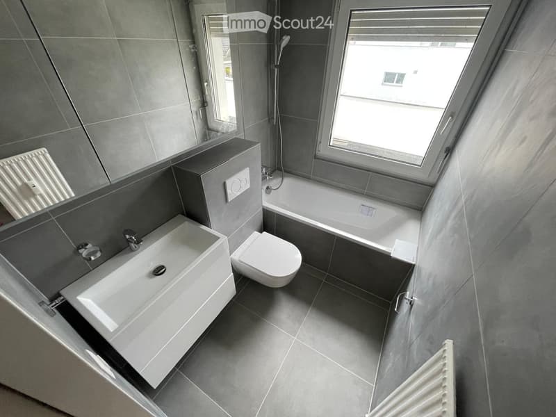 Modern bathroom with grey tiles, a white sink, toilet, bathtub, and a large mirror. Features a skylight window and radiator.