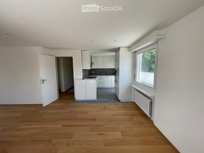 Empty room with wooden flooring, white walls, and a white kitchen area visible through an open doorway. The kitchen features white cabinets and a sink.