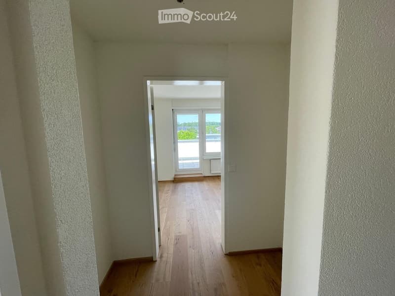 Empty hallway with wooden flooring and light-colored walls, leading to an open doorway that reveals a balcony area with a view.