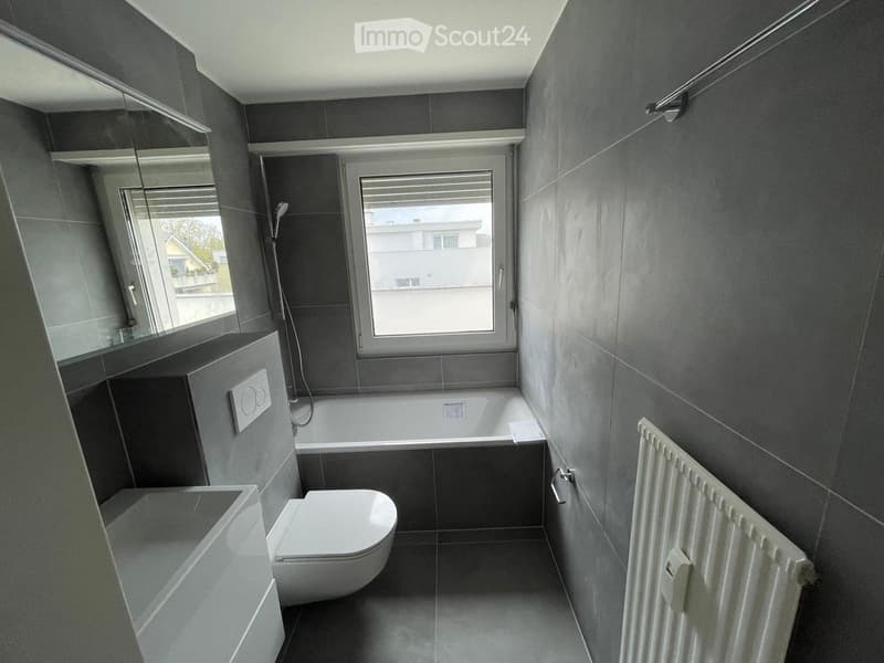 Modern bathroom with dark grey tiles, white toilet, bathtub, radiator, and large windows with blinds.