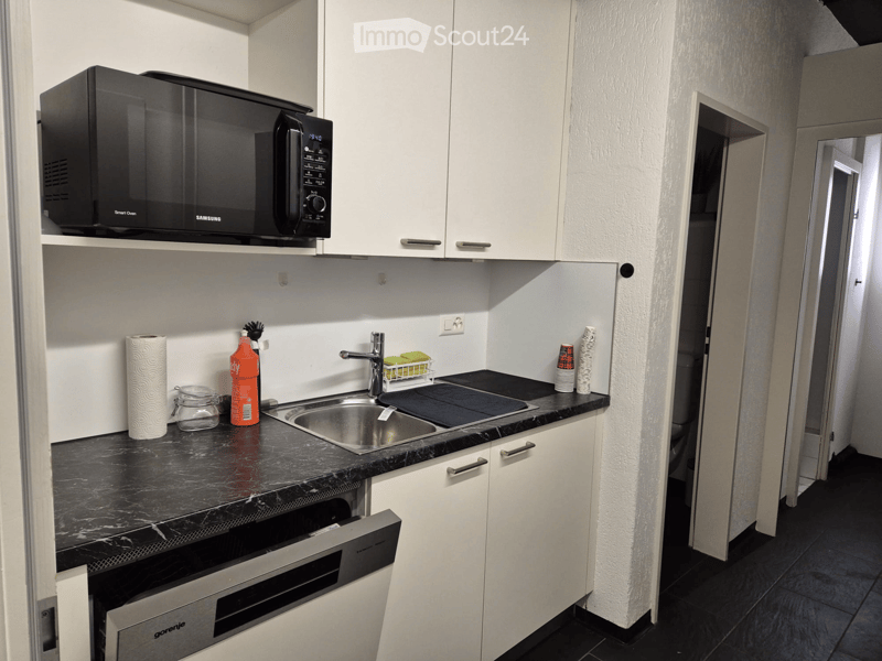 Modern kitchen with white cabinets, black countertop, stainless steel sink, microwave, and built-in oven. Items on the counter include a paper towel roll, bottle, cup, and jar.