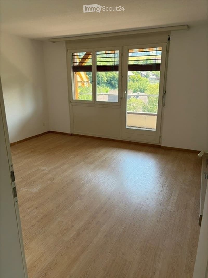 Empty room with wooden flooring, large windows with blinds, and a sliding door leading to a balcony.