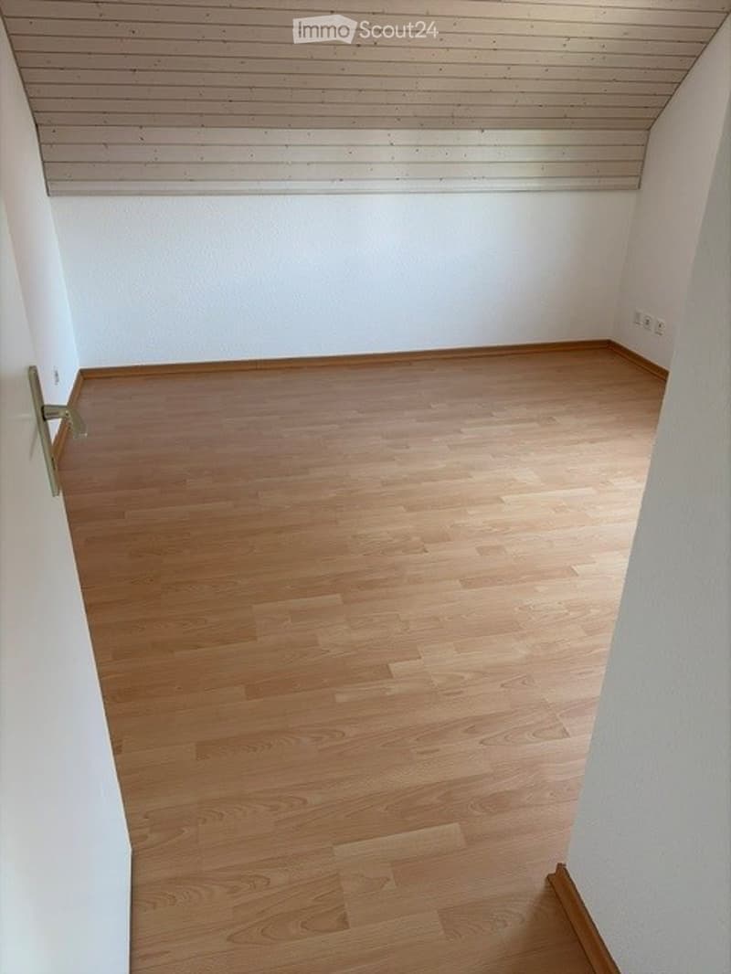 Empty room with light wooden flooring and white walls, featuring a simple wooden ceiling and a door frame visible on the left side.
