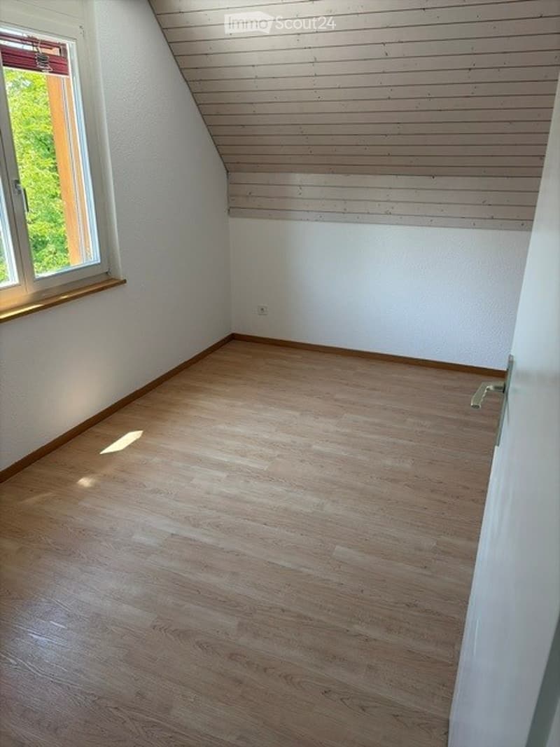 Completely empty room with wooden flooring, white walls, and a slanted wooden ceiling. Features a window with a view of greenery and a door.