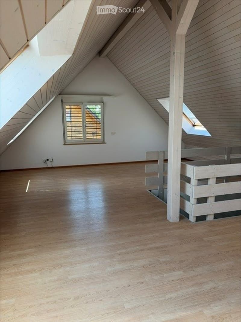 Bright, empty attic room with wooden flooring, white walls, and exposed wooden beams. Features a large window with shutters and a skylight, with a small wooden platform or landing area.