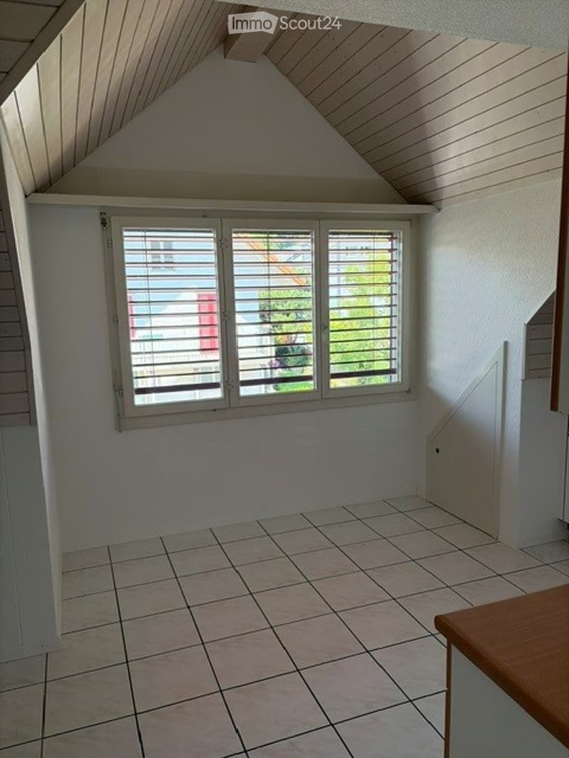 Empty room with white walls, tiled floor, and large window with blinds. Wooden ceiling beams and a small wooden table visible.
