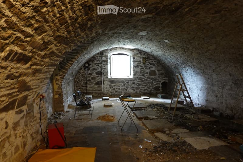 An unfinished room with stone walls and arched ceilings, featuring a single chair, a small table, and a ladder, with exposed construction materials and a window letting in natural light.