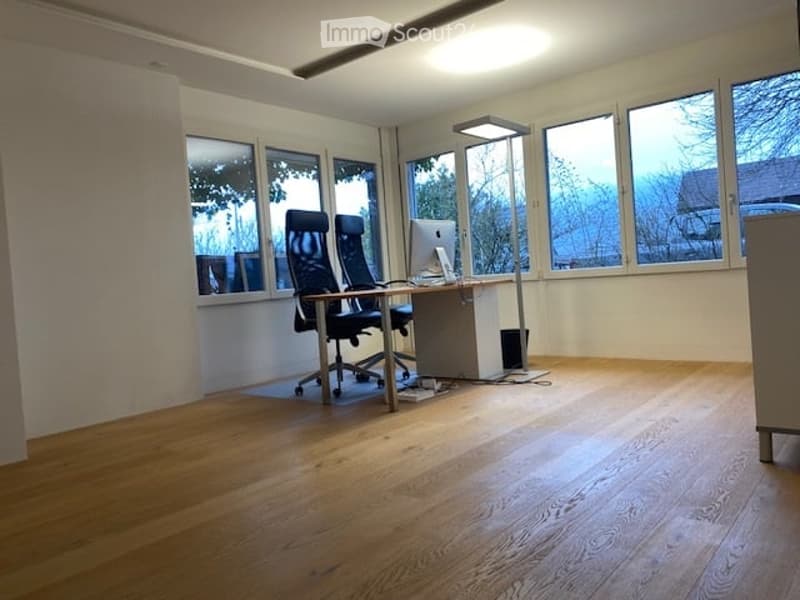 A large empty room with wooden floors, a desk with a computer, office chairs, large windows, and a ceiling lamp.