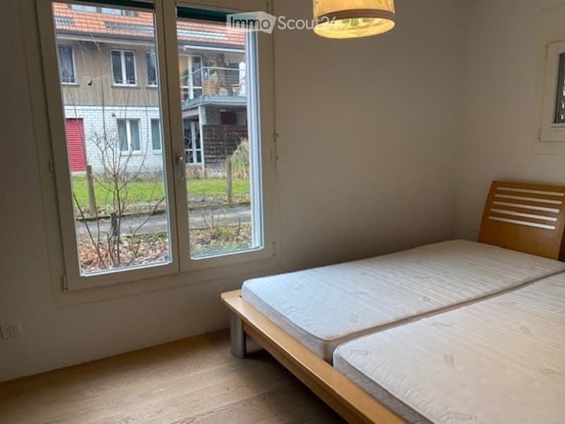 A bedroom with two beds, a large window, and a wooden headboard, overlooking a grassy lawn and a building with a balcony. Wooden floor and white walls.