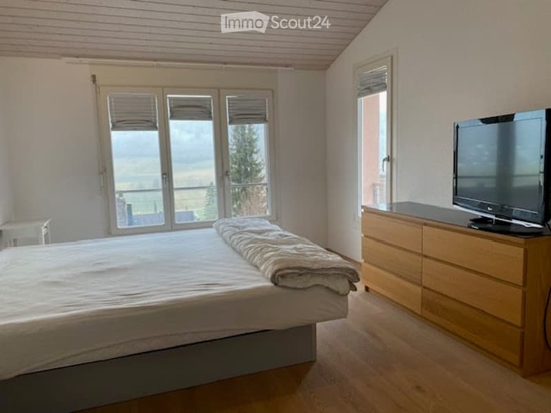 spacious bedroom with bed, TV, wooden dresser, large windows with views