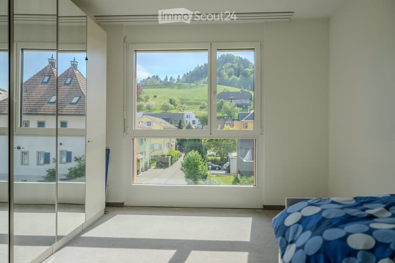 Bright bedroom with large windows offering scenic views of a village and countryside. Features a neatly made bed with a blue and white patterned duvet, a large mirror, and a carpeted floor.