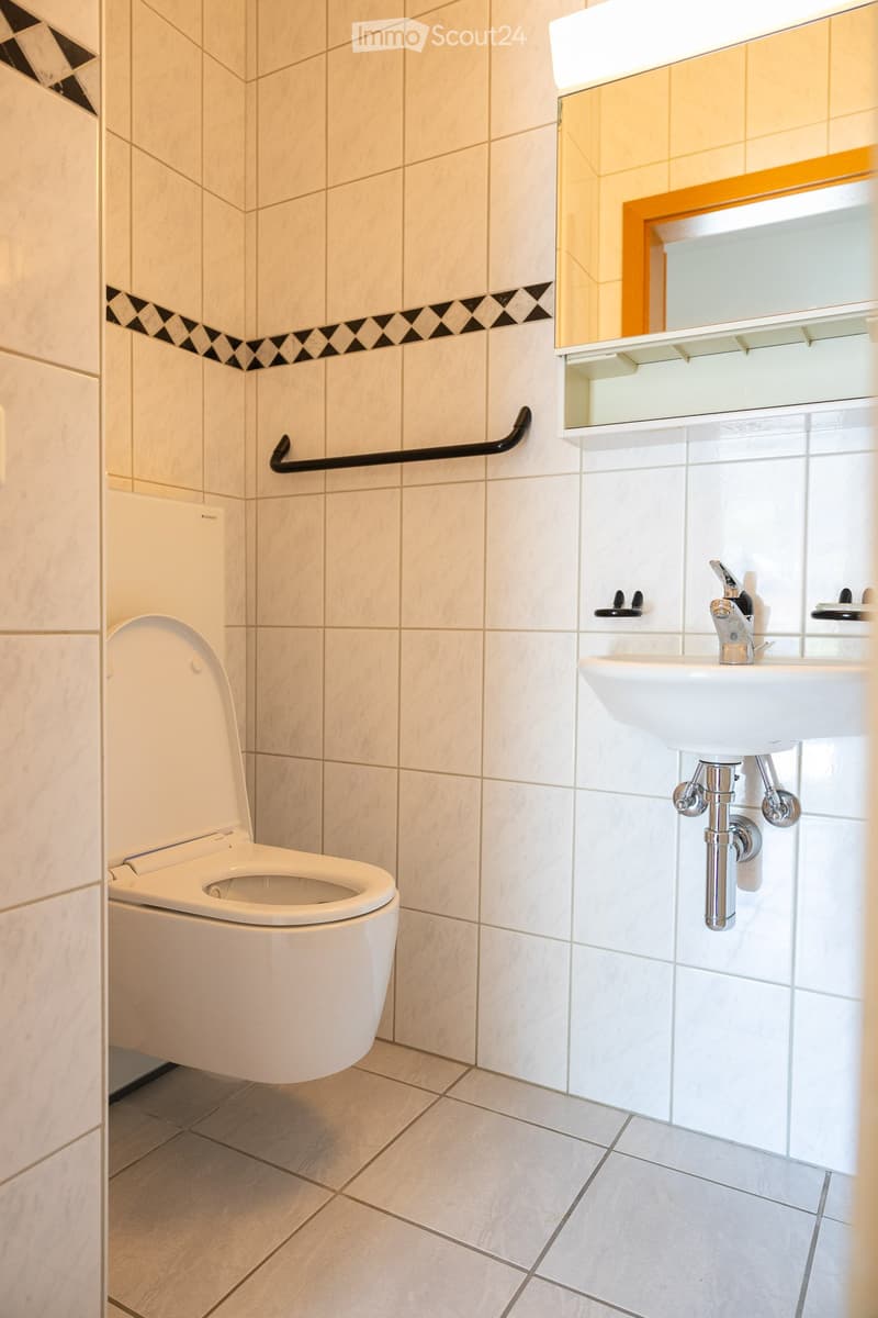 Modern bathroom with beige tiled walls, white toilet, white sink with chrome faucet, mirror with wooden frame, and a black handrail.