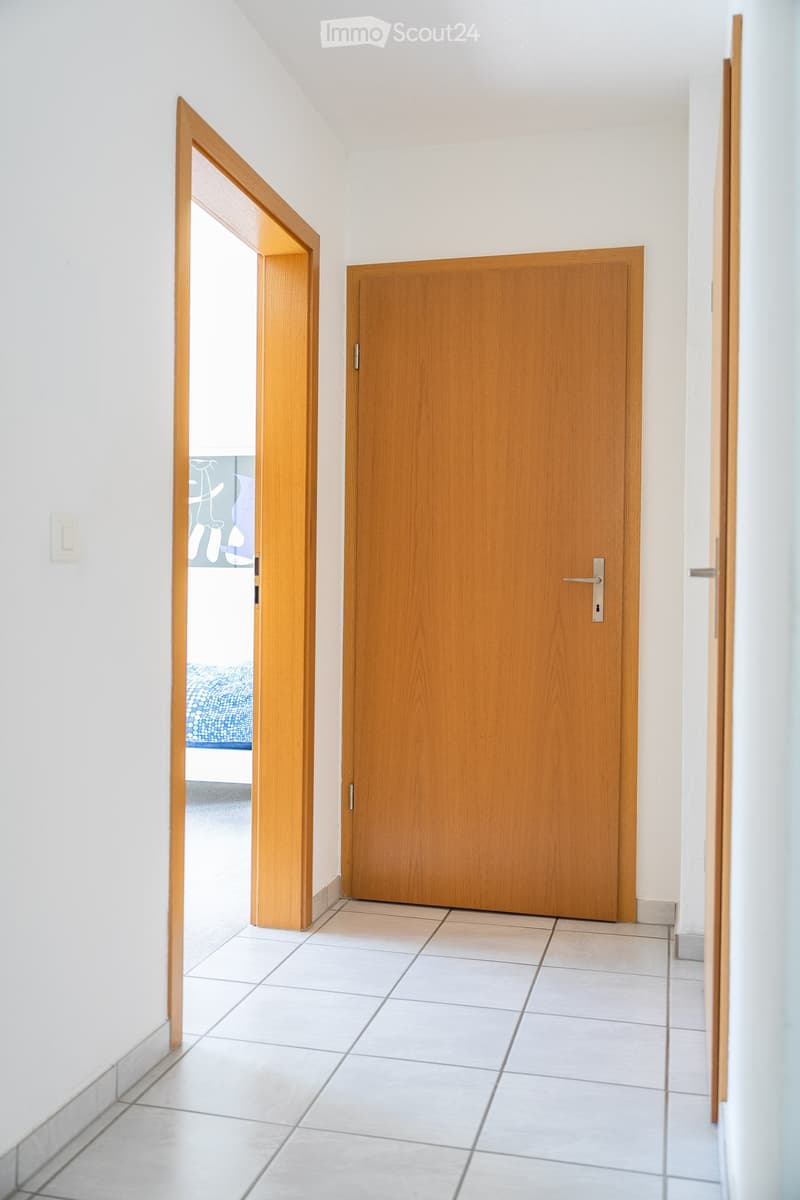 A clean, modern hallway with white tiled flooring, light-colored walls, and wooden doors. One door is open, revealing a glimpse of a bedroom with a blue bedspread.