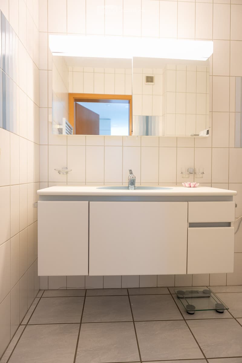 Modern bathroom with white tiled walls and floor, a white sink cabinet with a chrome faucet, a large mirror above the sink, and a glass scale on the floor.