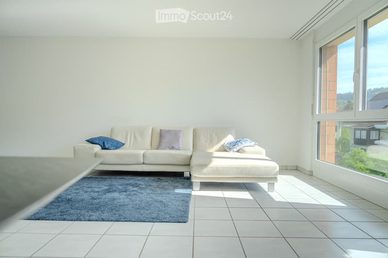 Bright living room with white walls, tiled floor, large window offering a scenic view, a white sectional sofa with an ottoman, and a blue rug.