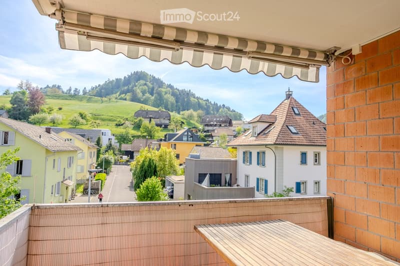 Balcony with wooden table and cushioned seating, overlooking a scenic town with houses, greenery, and hills.