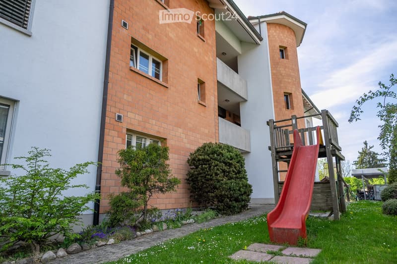 A well-maintained garden area with a red slide, small trees, shrubs, and a paved pathway leading to the building entrance.