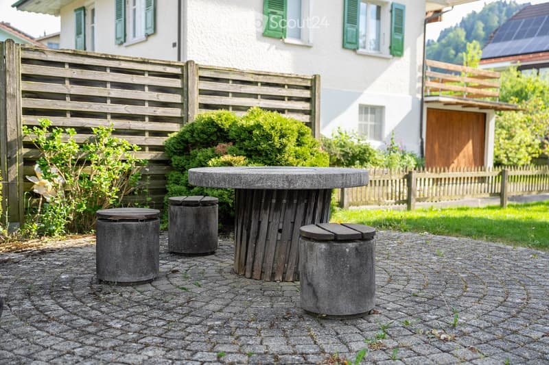 Garden area with a round stone table and four stone stools on a cobblestone patio, surrounded by greenery and a wooden fence.