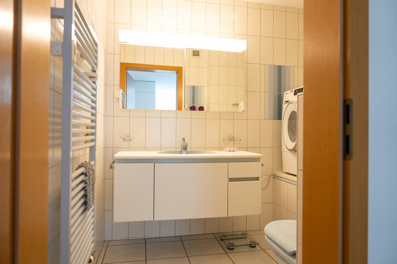 Modern bathroom with white tiled walls, a sink with a modern faucet, a large mirror, a toilet, a washing machine, and a radiator.