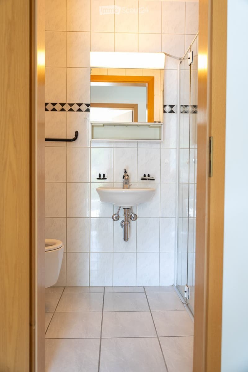 Modern bathroom with white tiles, a sink with a modern faucet, a toilet, a mirror with a wooden frame, and a glass-enclosed shower area.