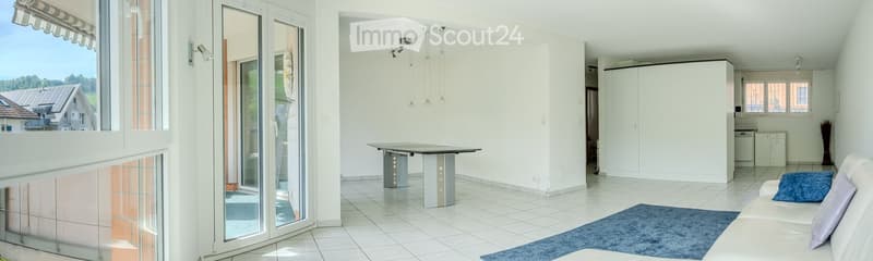 Bright living room with white walls, tiled floor, large windows, and a view of the countryside. Features a white sofa, a blue throw pillow, a modern table, and built-in white cabinets.