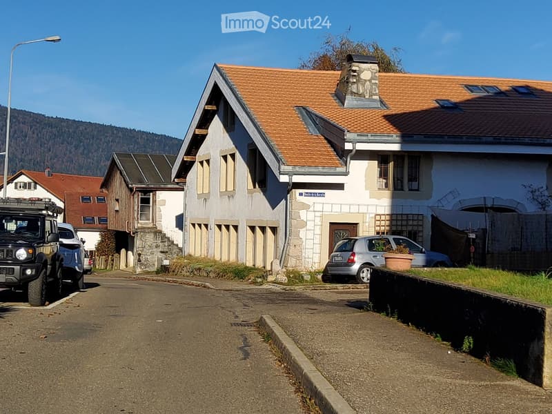 Two-story house with a red-tiled roof, white walls, and multiple windows. A stone chimney is visible on the roof. The house has a small balcony and a stone staircase leading to the entrance. There are several parked cars in front of the house, and the sur