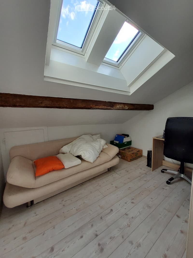 Cozy attic room with wooden flooring, skylight windows, a small sofa with pillows, a desk, and a chair.