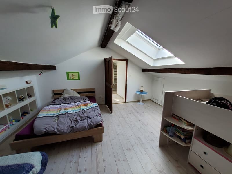 Cozy attic bedroom with wooden floors, slanted ceilings, skylight windows, and built-in storage. Features a single bed with colorful bedding, a small round table with a lamp, and shelves with books and toys.