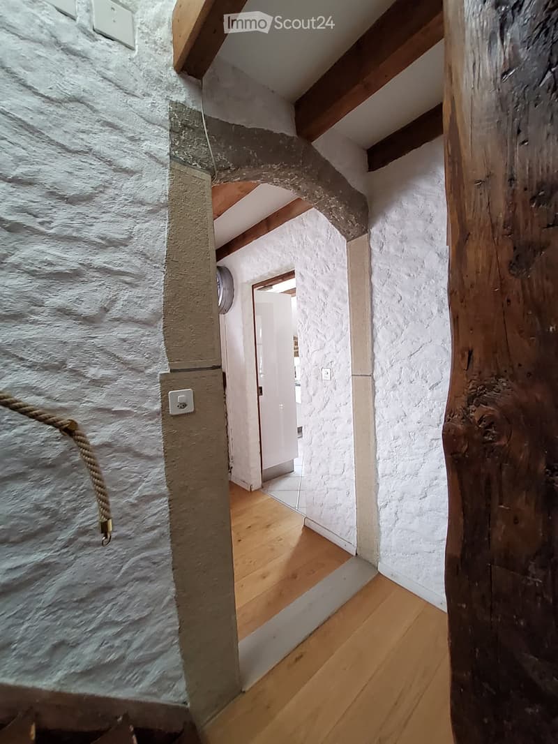 A narrow hallway with wooden flooring, white plaster walls, and exposed wooden beams on the ceiling. A rope handrail is visible on the left side, and a doorway leads to another room.