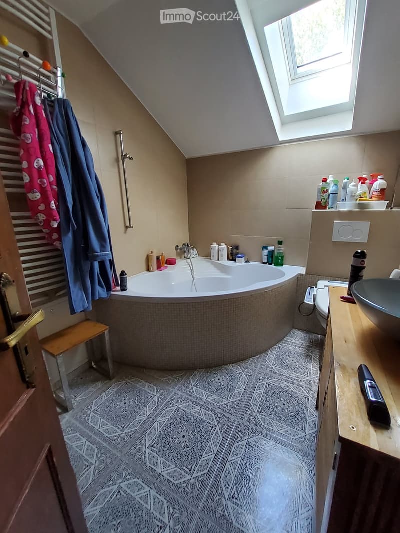 Bathroom with a bathtub, sink, toilet, and various toiletries. Features a wooden stool, towels, and a patterned tile floor.