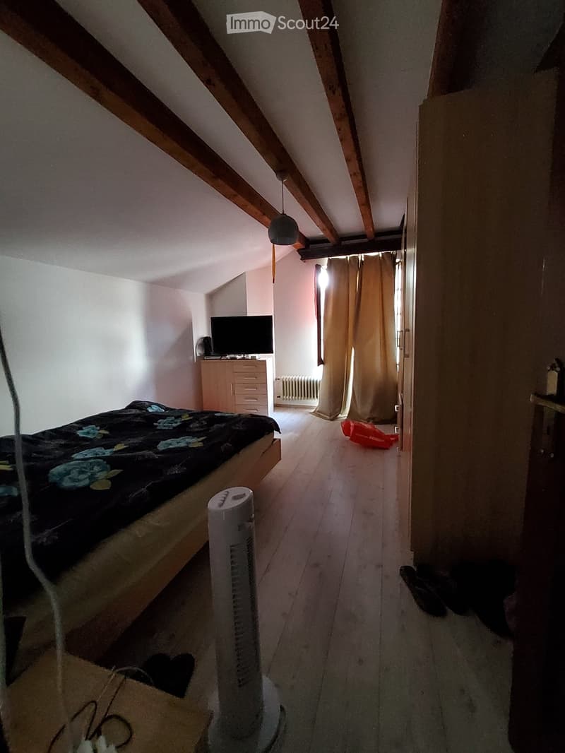 Bedroom with slanted wooden ceiling beams, wooden floor, bed with floral bedding, fan, television on dresser, radiator, and open closet area.
