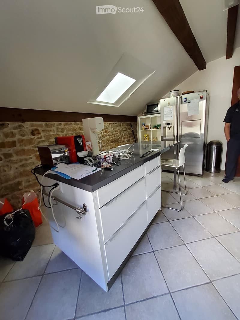 Modern kitchen with white cabinets, stainless steel refrigerator, microwave, and a small seating area with a chair and stool. The kitchen features a stone accent wall and a skylight above the counter.