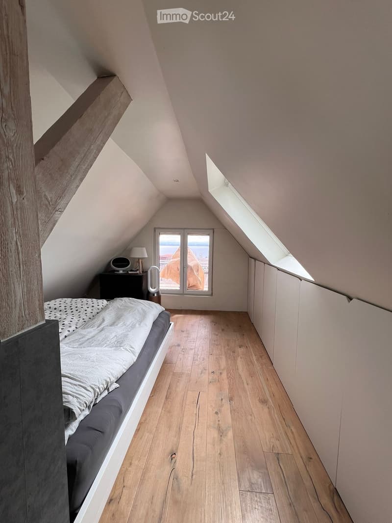 Cozy attic bedroom with wooden flooring, slanted ceilings, and large windows offering a view. Features a single bed with bedding, a nightstand with a lamp, and a speaker on the nightstand.
