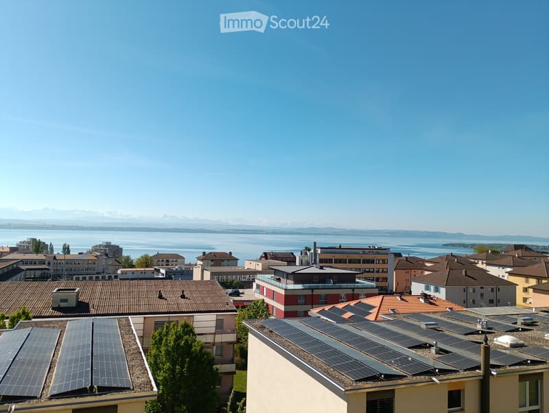 A panoramic view of a residential area with multiple buildings, solar panels on rooftops, and a scenic view of a lake and distant mountains under a clear blue sky.