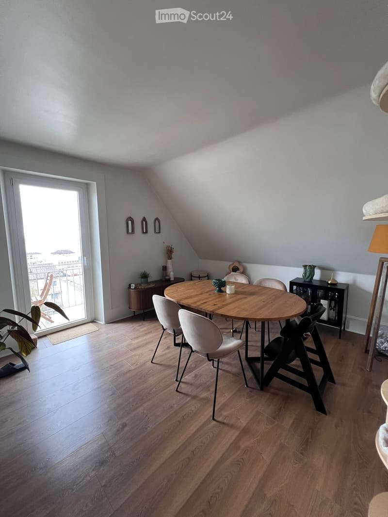 Bright, modern dining area with wooden flooring, round wooden dining table surrounded by white chairs, small side table, decorative vases, and a view through a large window.