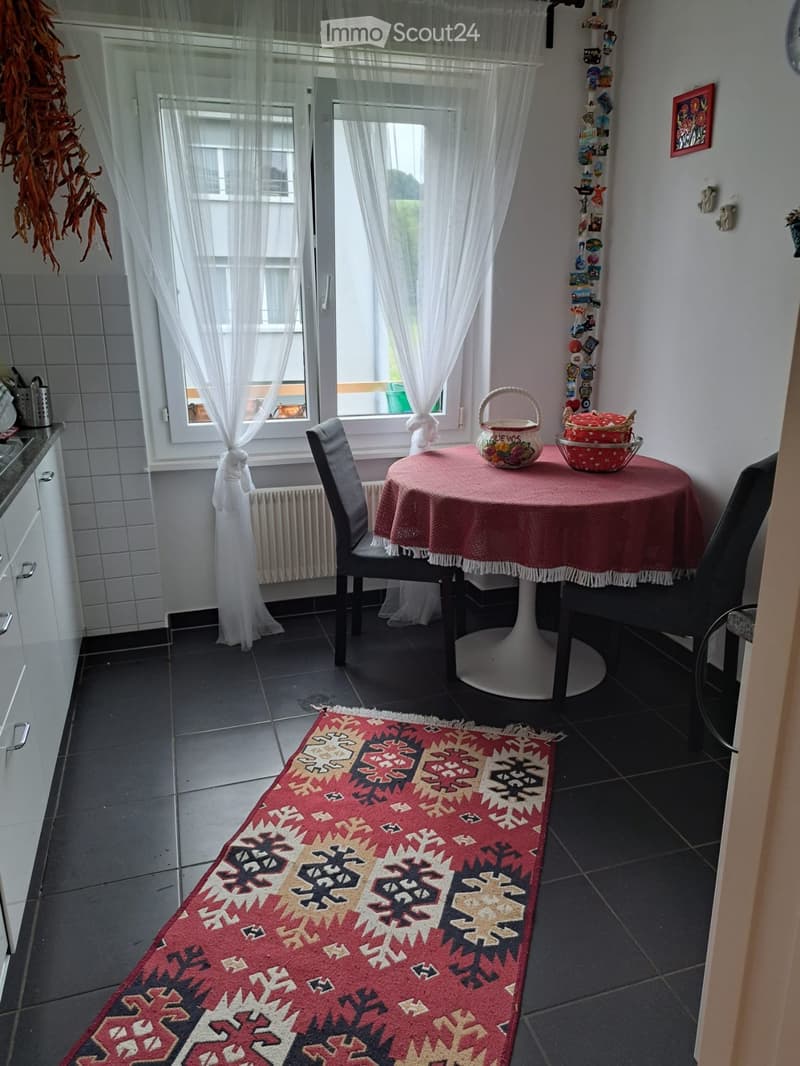 A small dining area with a round table, two chairs, a rug, and a window with curtains. The table has a red tablecloth and a basket on top. The room has a white wall, a radiator, and a picture frame. The floor is tiled, and there is a cabinet with drawers