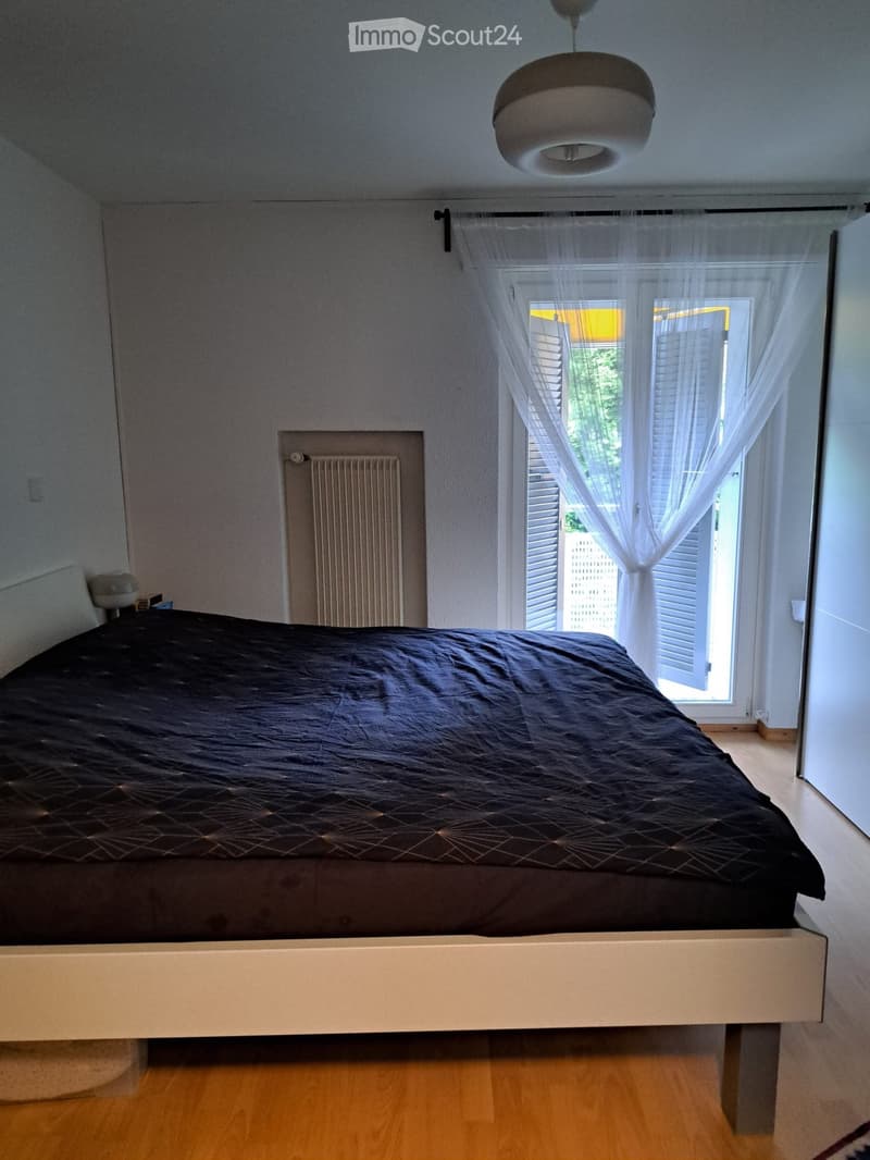 Bedroom with bed, white walls, wooden floor, white curtains, white radiator, lamp, window with shutters